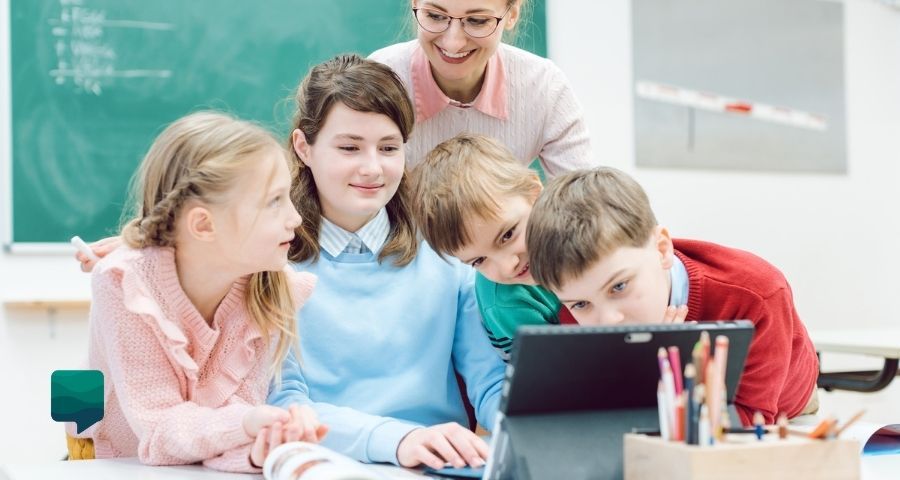 Tecnologia no ensino - foto de 4 crianças brancas em frente a um computador enquanto a professora está atrás deles sorrindo.